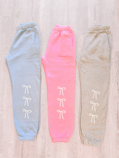 Light blue, pink, and gray sweatpants with white bow details, displayed on a wood background.
