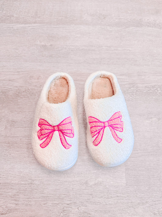 Cozy white slippers with pink bow embroidery, shown on light wood floor.