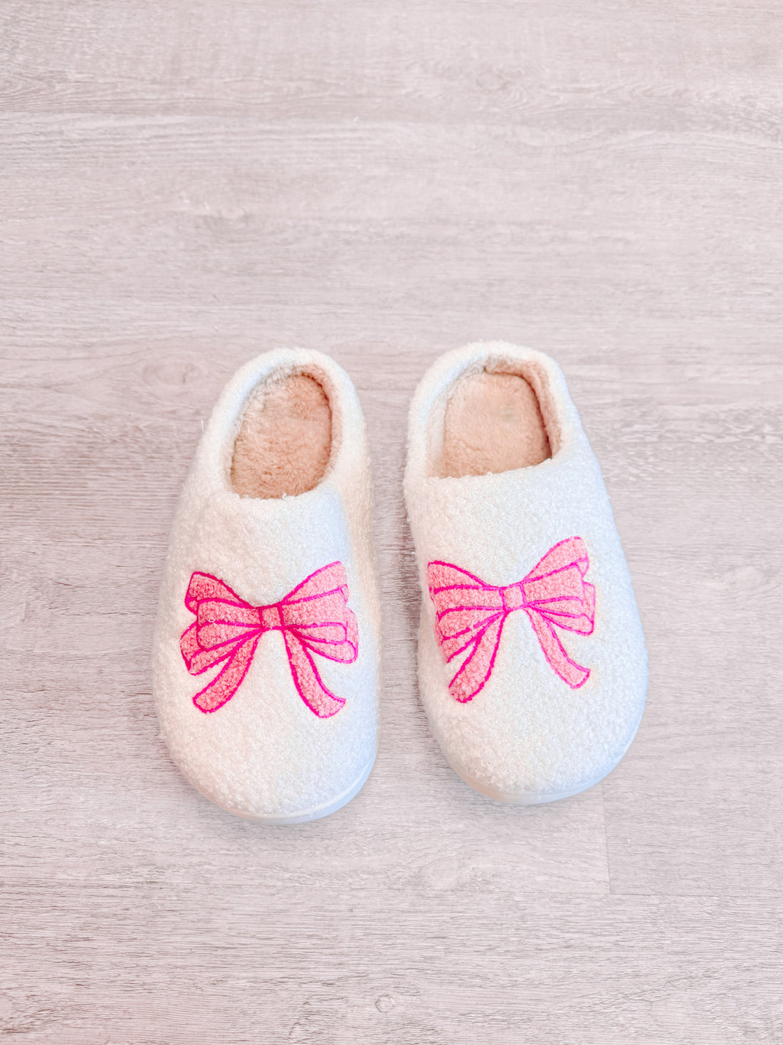 Cozy white slippers with pink bow embroidery, shown on light wood floor.
