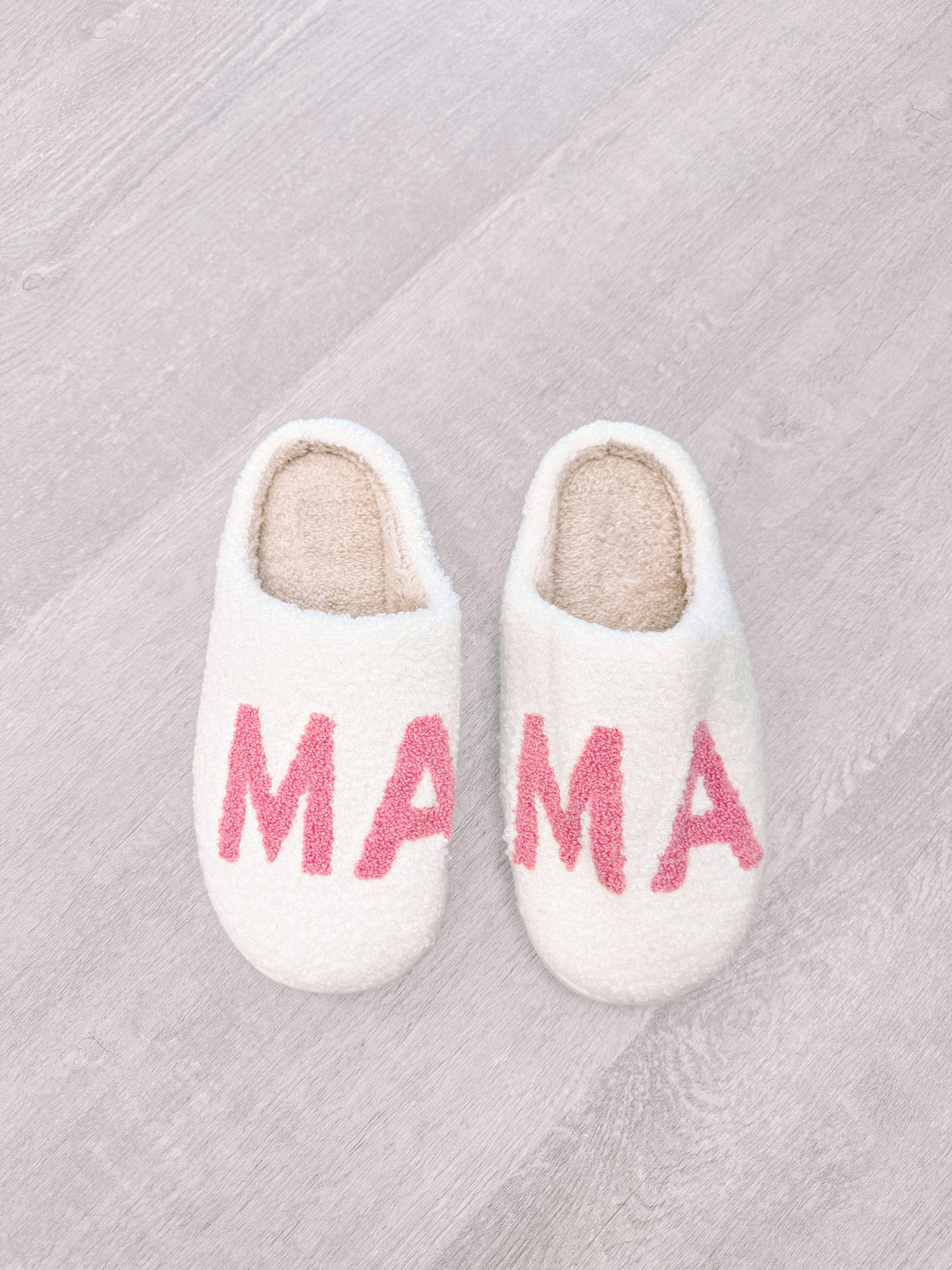 White fluffy slippers with pink "Mama" lettering, shown on a wood floor.
