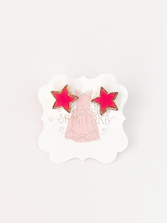 Pink star-shaped earrings with gold accents, displayed on a card.
