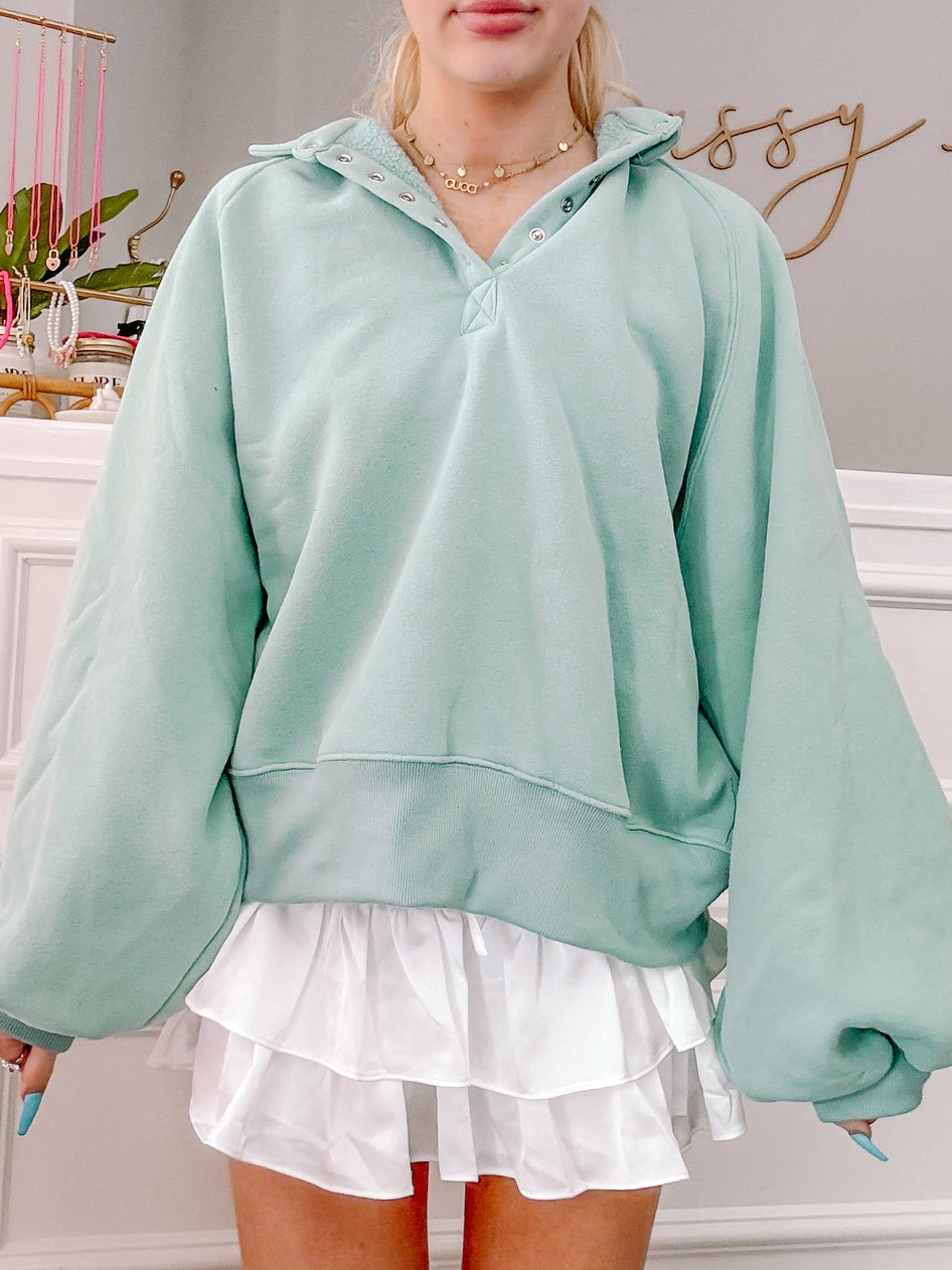 Mint green pullover sweatshirt with balloon sleeves and a buttoned henley neckline, paired with a white ruffled mini skirt.
