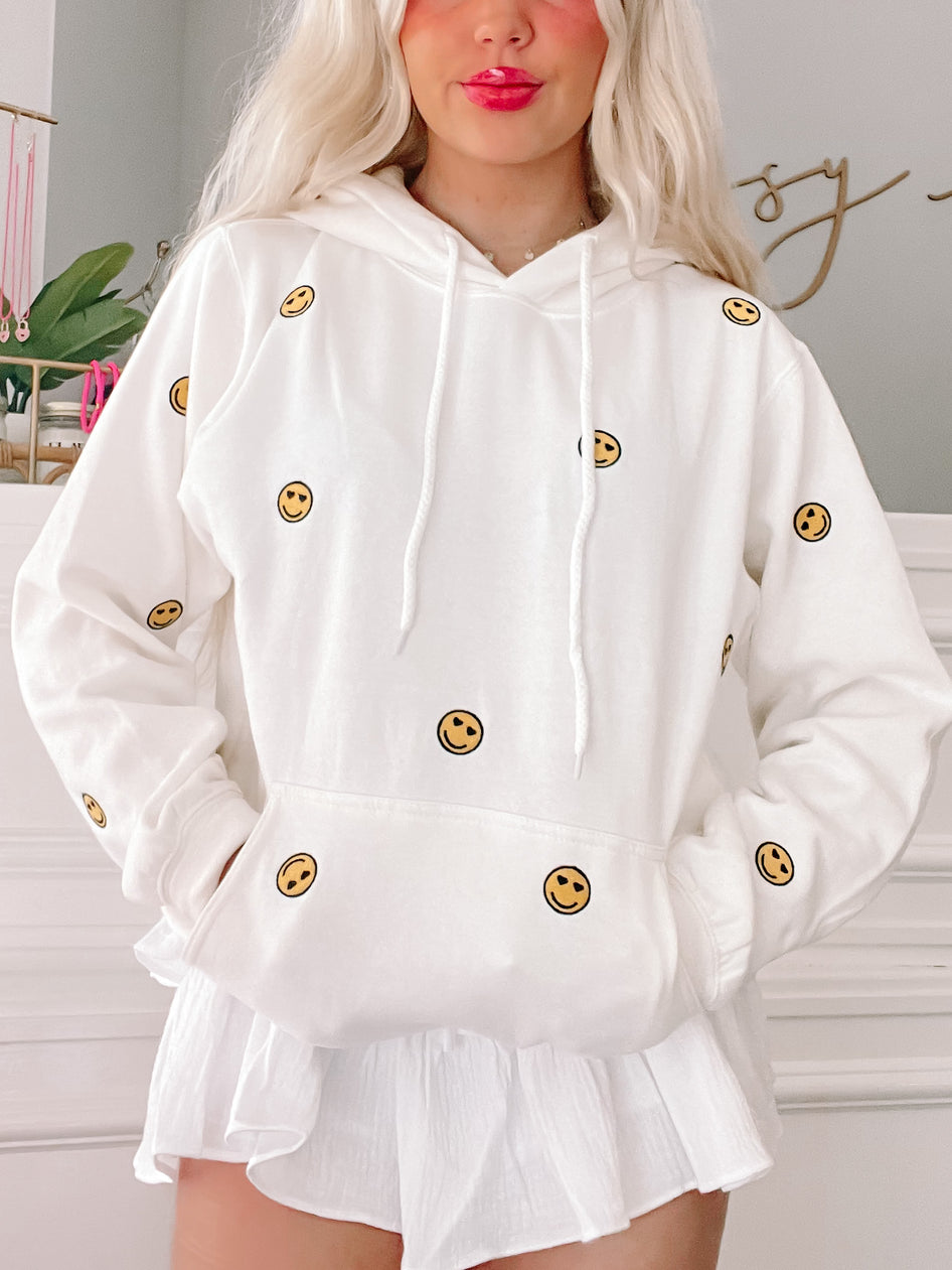 White hoodie featuring small, embroidered smiley faces, shown on a model.
