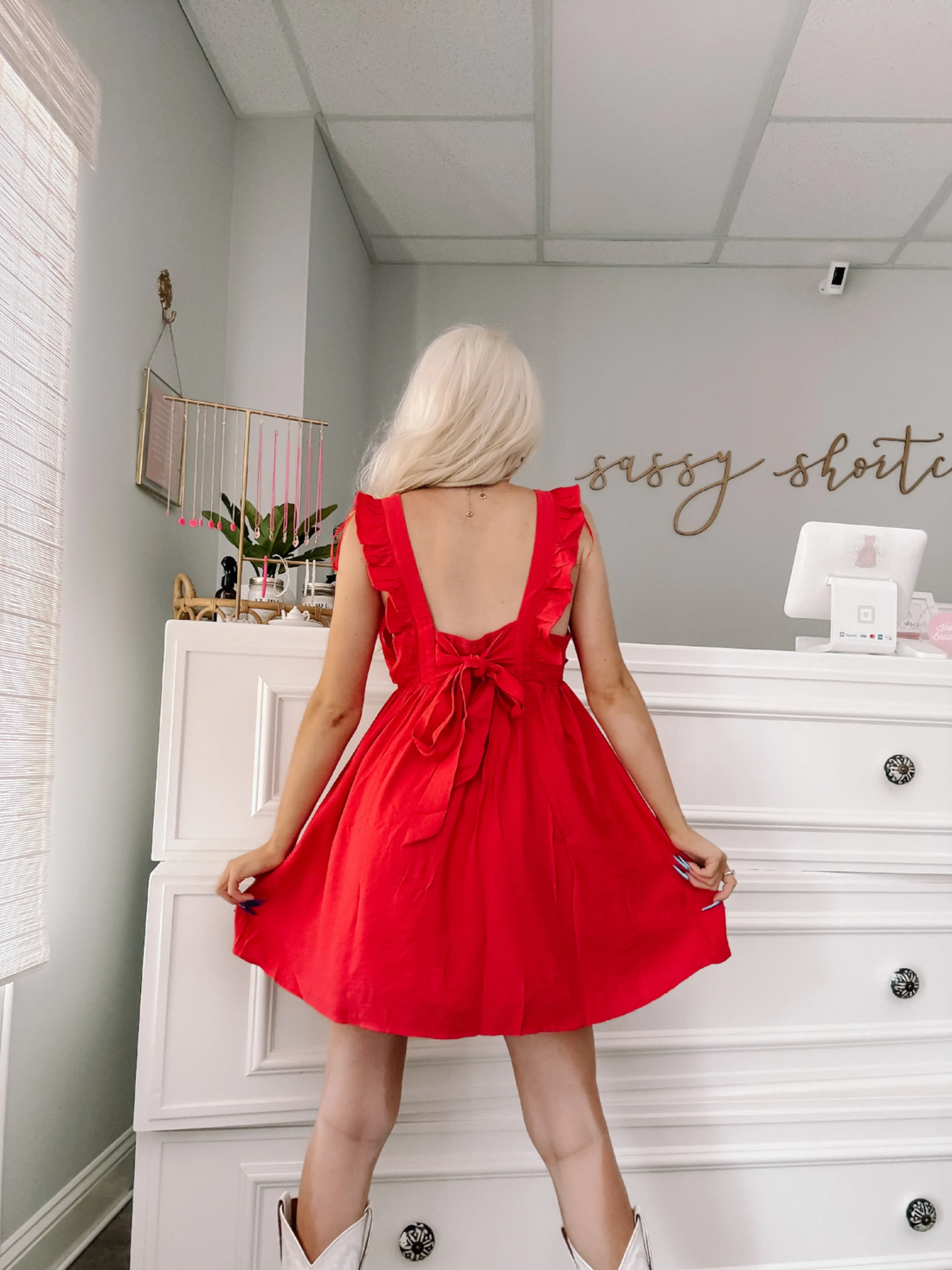 Red ruffle mini dress with a back bow detail, shown from behind in a bright room.
