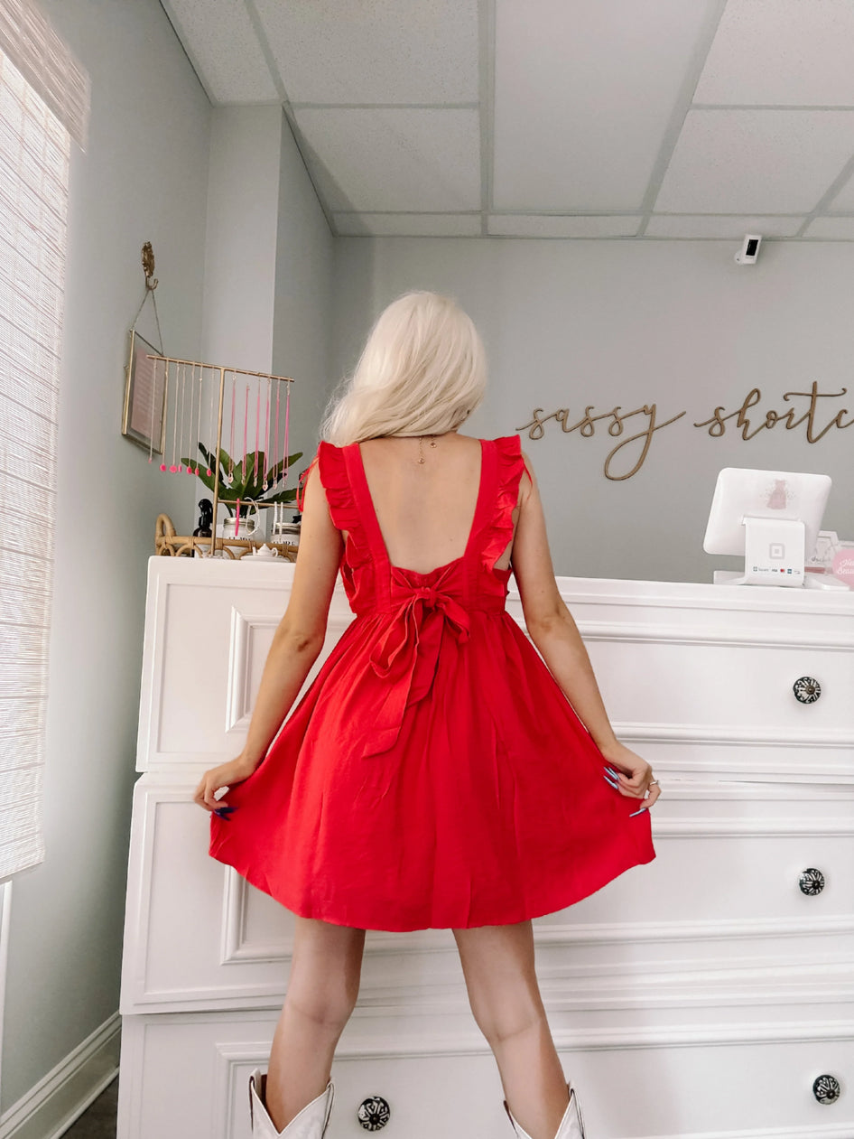 Red ruffle mini dress with a back bow detail, shown from behind in a bright room.
