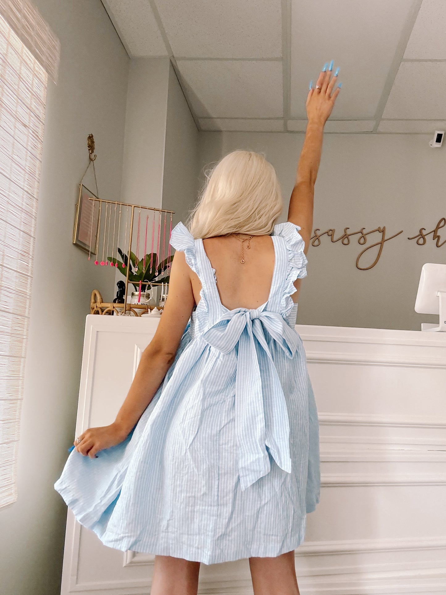 Light blue and white striped dress with ruffle details and bow back, shown on model in a bright room.
