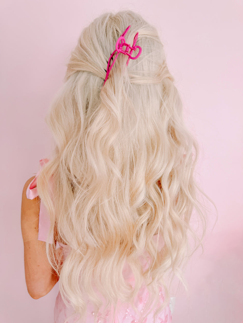 Pink claw hair clip in blonde wavy hair against a pink background.
