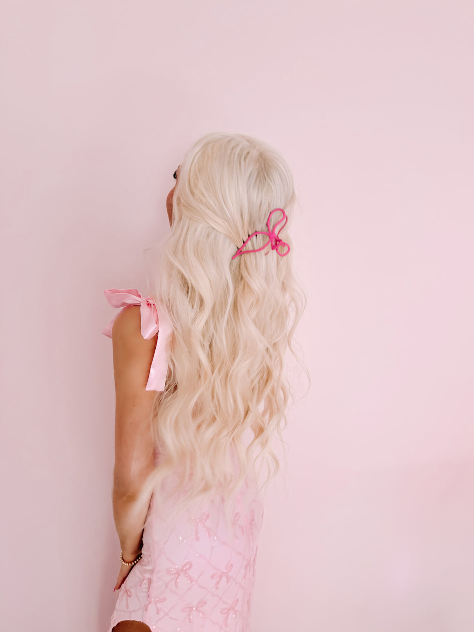 Pink bow hair clip in blonde hair against a pink background.
