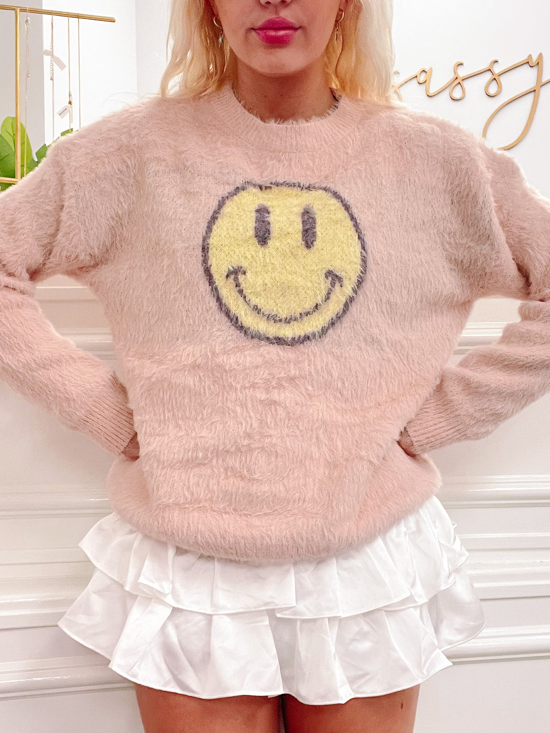 Fuzzy pink sweater featuring a playful yellow smiley face, styled with a white ruffle skirt.
