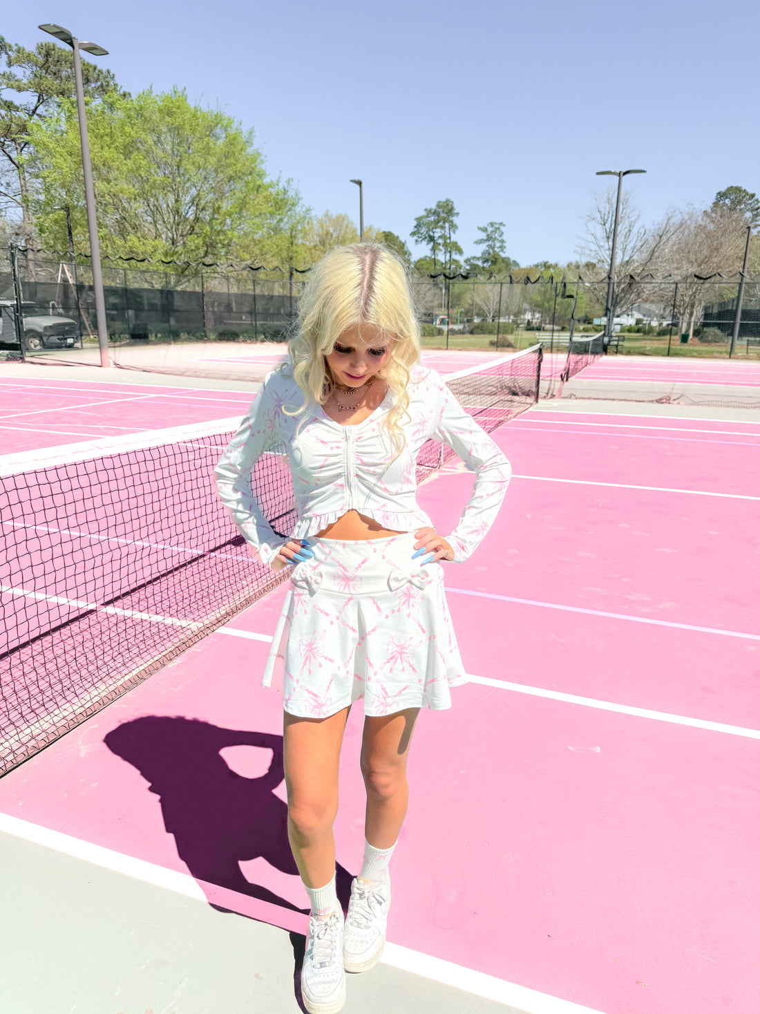 Model wearing a white bow-print skirt and matching top on a pink tennis court.
