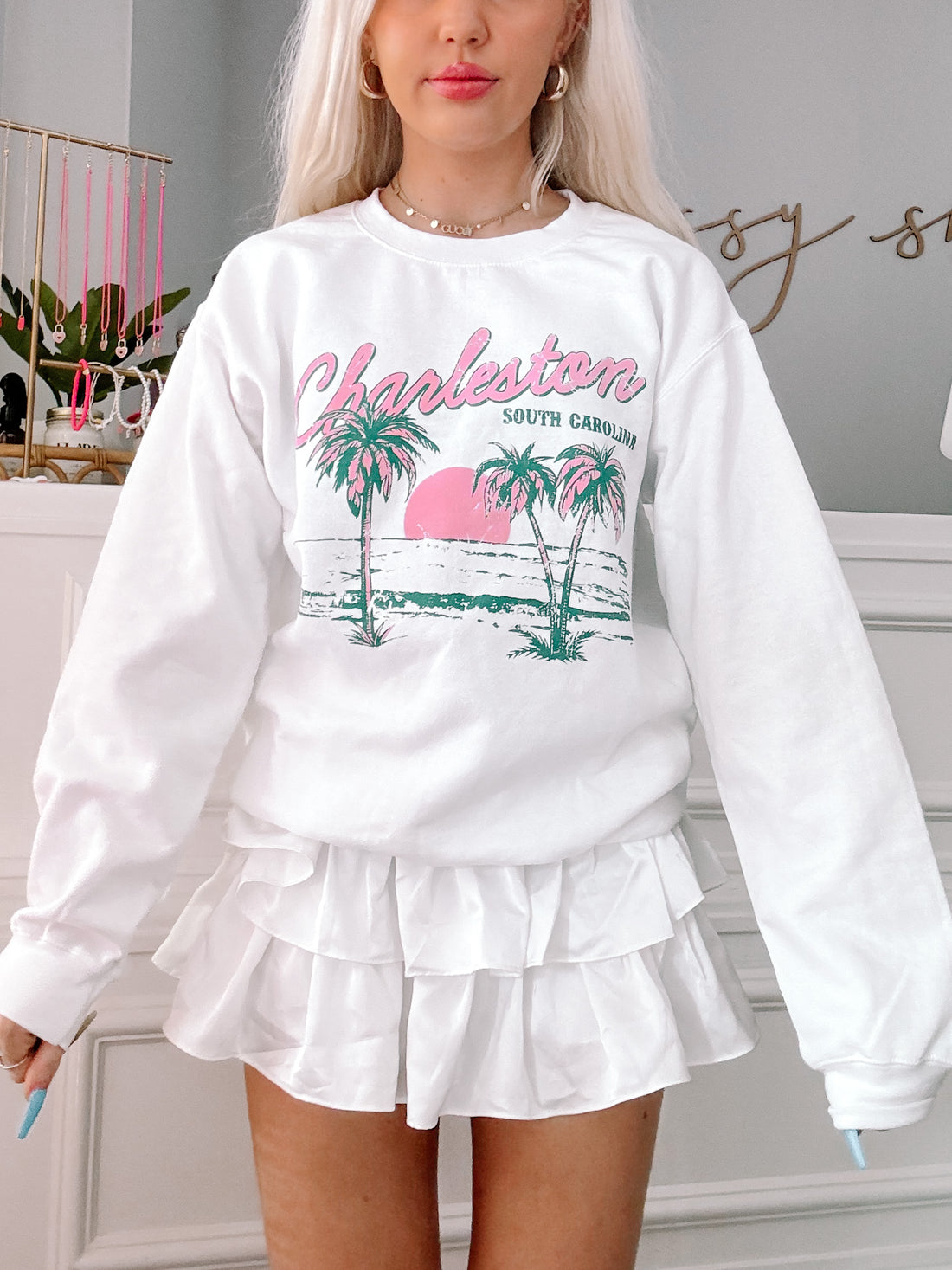 White crewneck sweatshirt featuring a palm tree and sunset graphic, paired with a white ruffle skirt.
