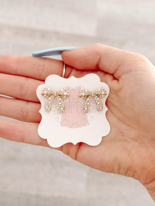 Pearl and gold bow earrings displayed on a card in a hand.
