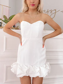 White strapless mini dress with a ruffled hem, shown on a model against a white wall.
