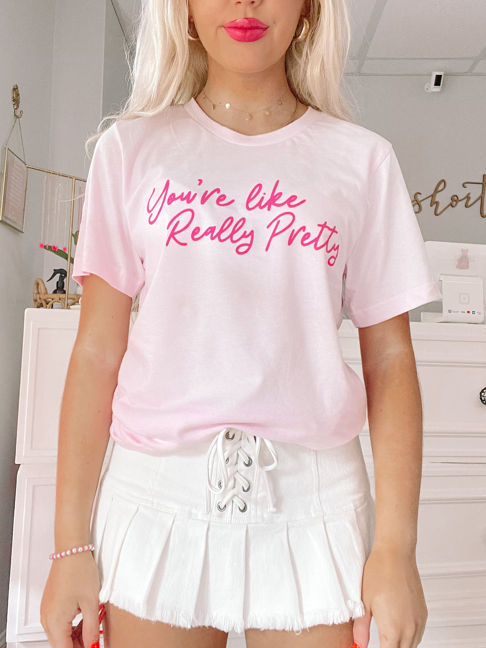 Pink t-shirt featuring a playful slogan, styled with a white pleated skirt.
