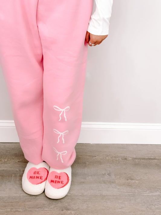 Pink sweatpants with delicate bow details, paired with heart-shaped slippers; cozy Valentine's Day style.