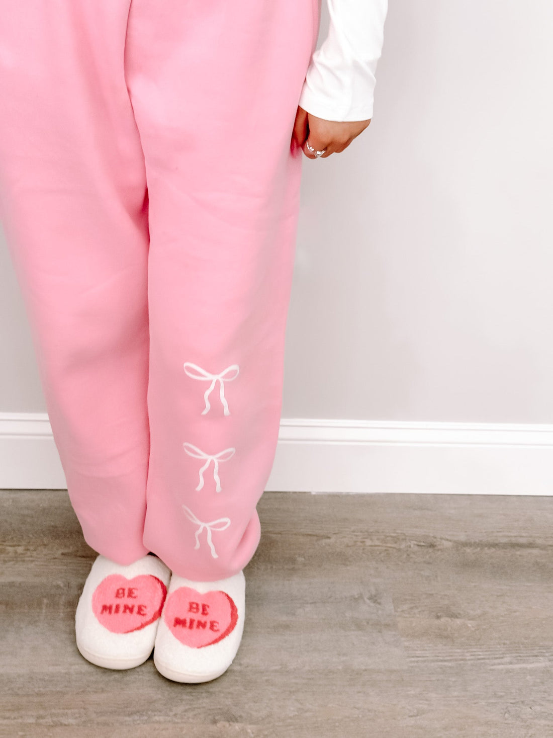 Pink sweatpants with bow details, paired with "Be Mine" slippers; shown against a light wood floor.
