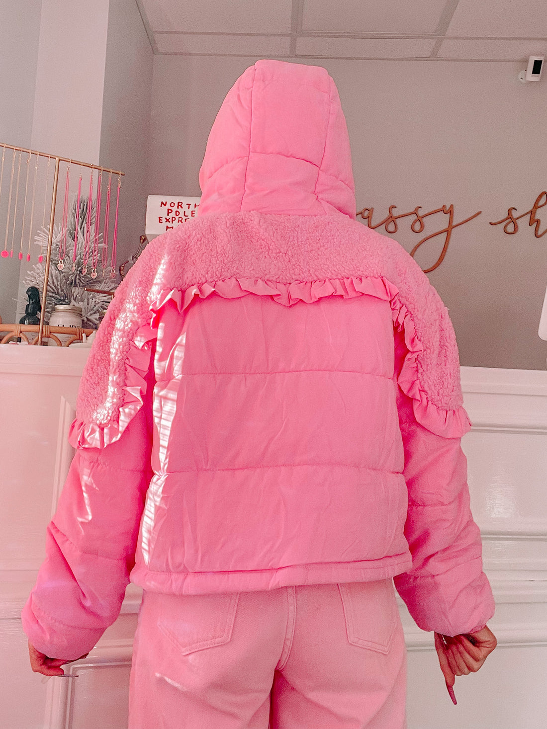 Pink puffer jacket with sherpa trim and ruffle details, shown from the back in a bright interior.
