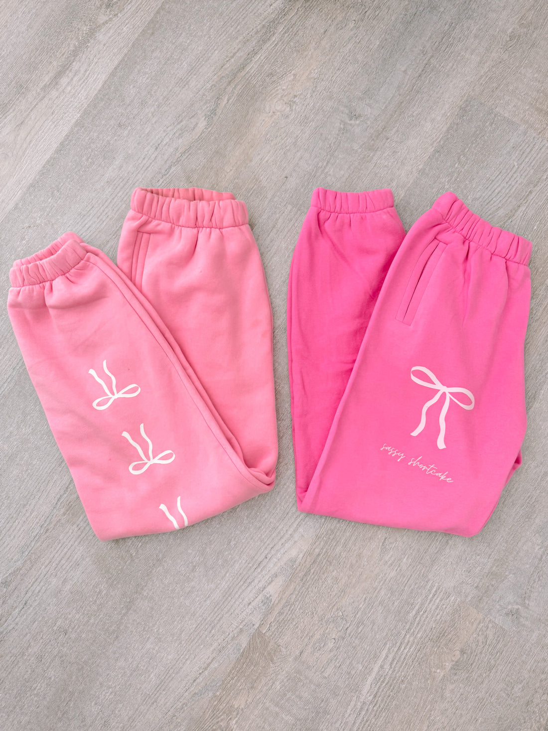 Pink sweatpants with bow details, flat lay on wood.
