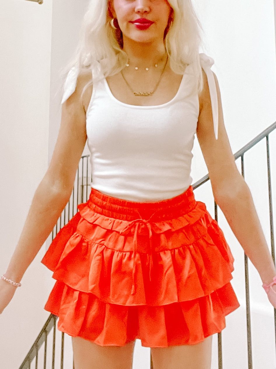 Bright orange tiered ruffle mini skirt with drawstring waist, modeled indoors near a staircase.

