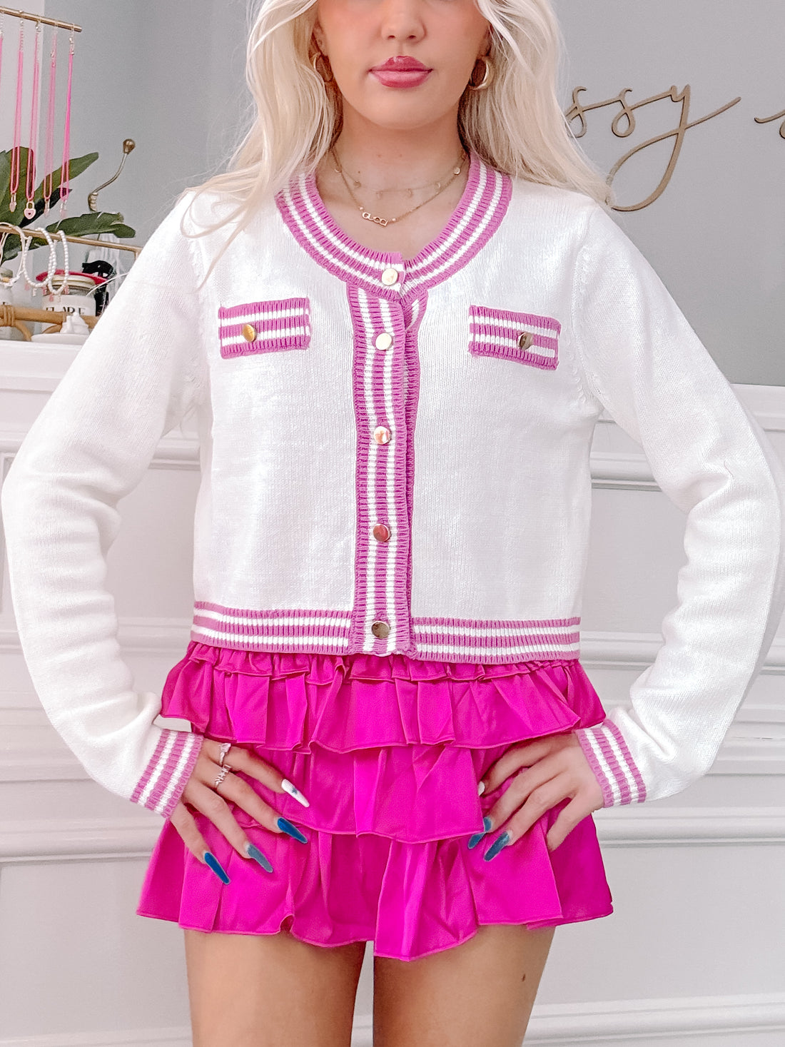 White cropped cardigan with purple trim, button closure, and gold buttons, modeled with a fuchsia skirt.
