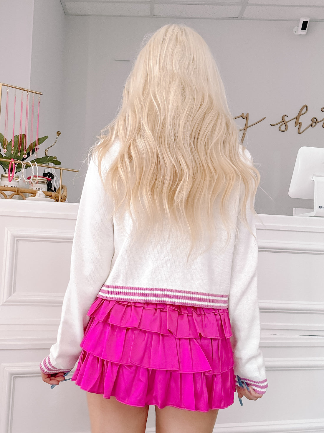 White cropped sweater with purple trim, shown from the back paired with a bright pink ruffled skirt.
