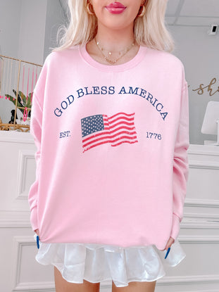 Pink crewneck sweatshirt with an American flag graphic and "God Bless America, Est. 1776" text.
