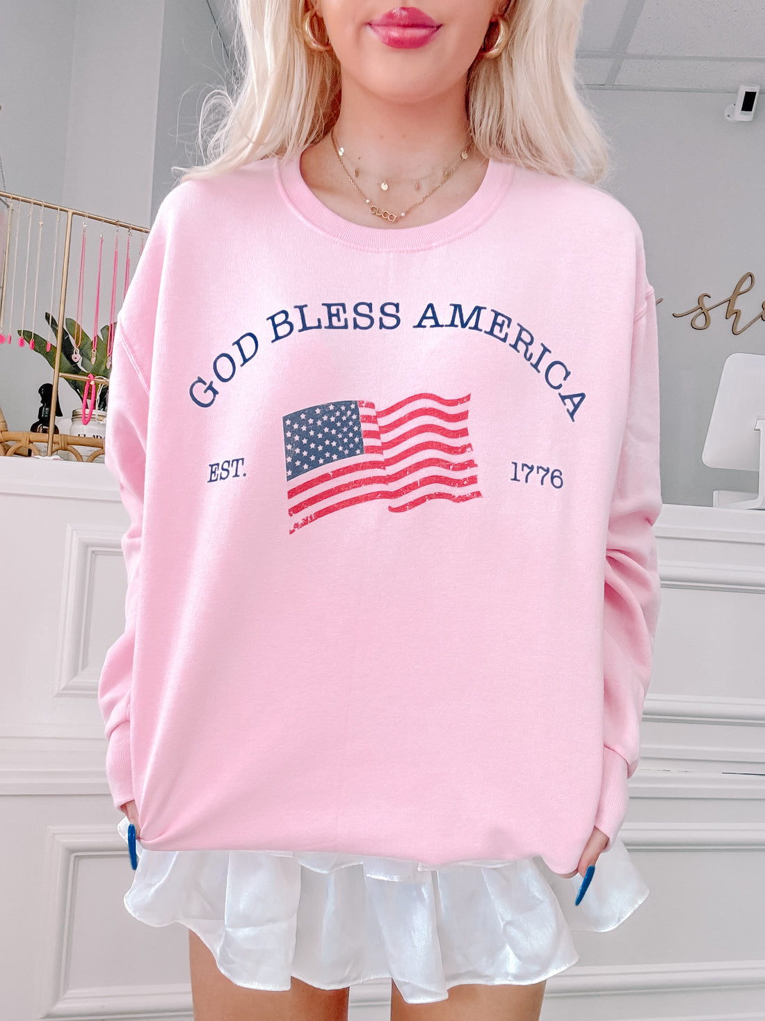 Pink crewneck sweatshirt with an American flag graphic and "God Bless America, Est. 1776" text.
