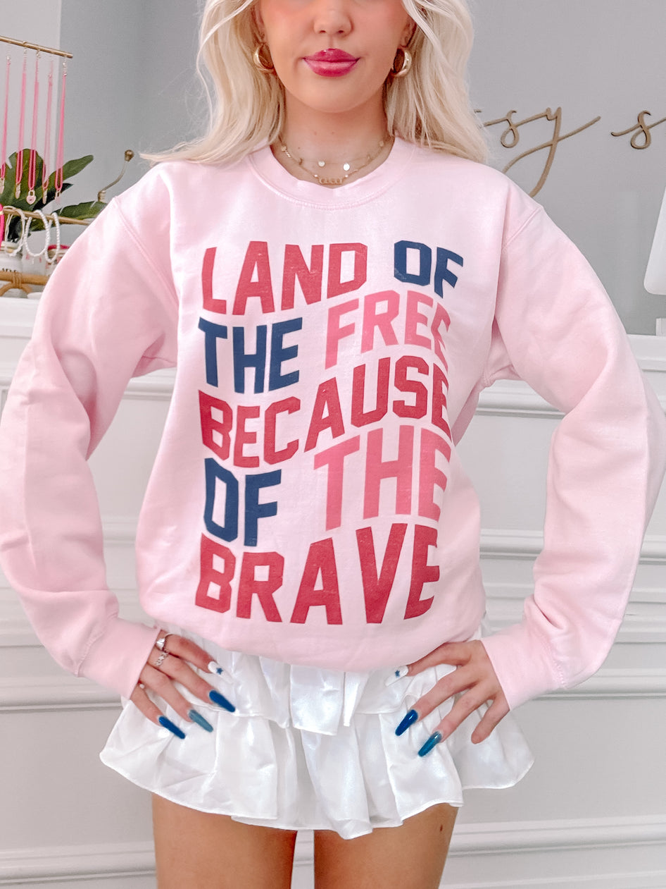Pink crewneck sweatshirt featuring a patriotic slogan, "Land of the Free Because of the Brave," in red, navy, and pink lettering.
