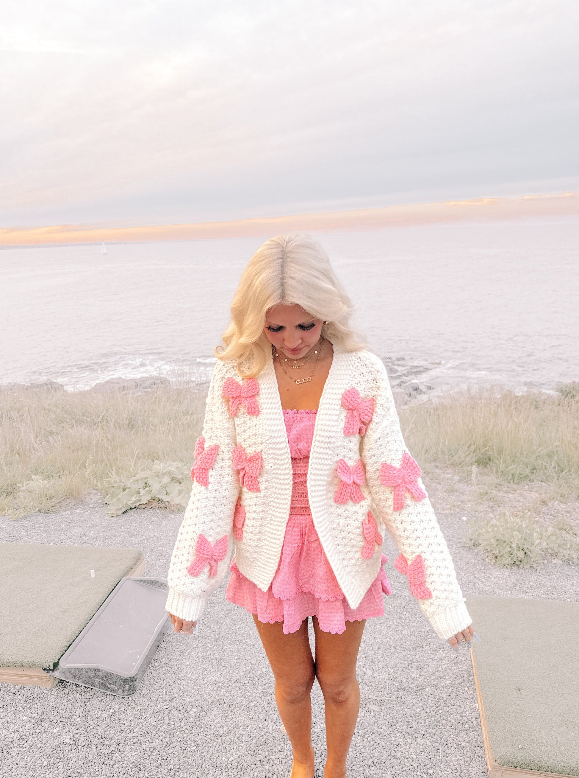 Woman in white knit cardigan adorned with pink bows, posing outdoors by the sea.
