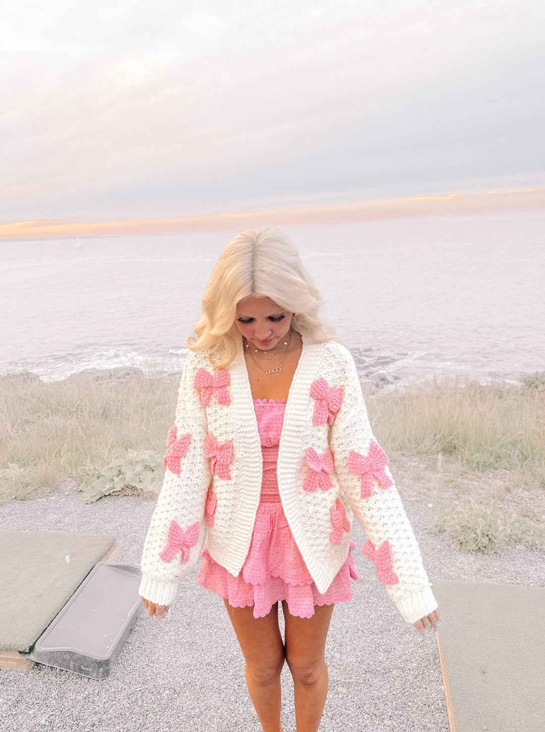 Pink and white knit cardigan with bow details, worn outdoors near the ocean.
