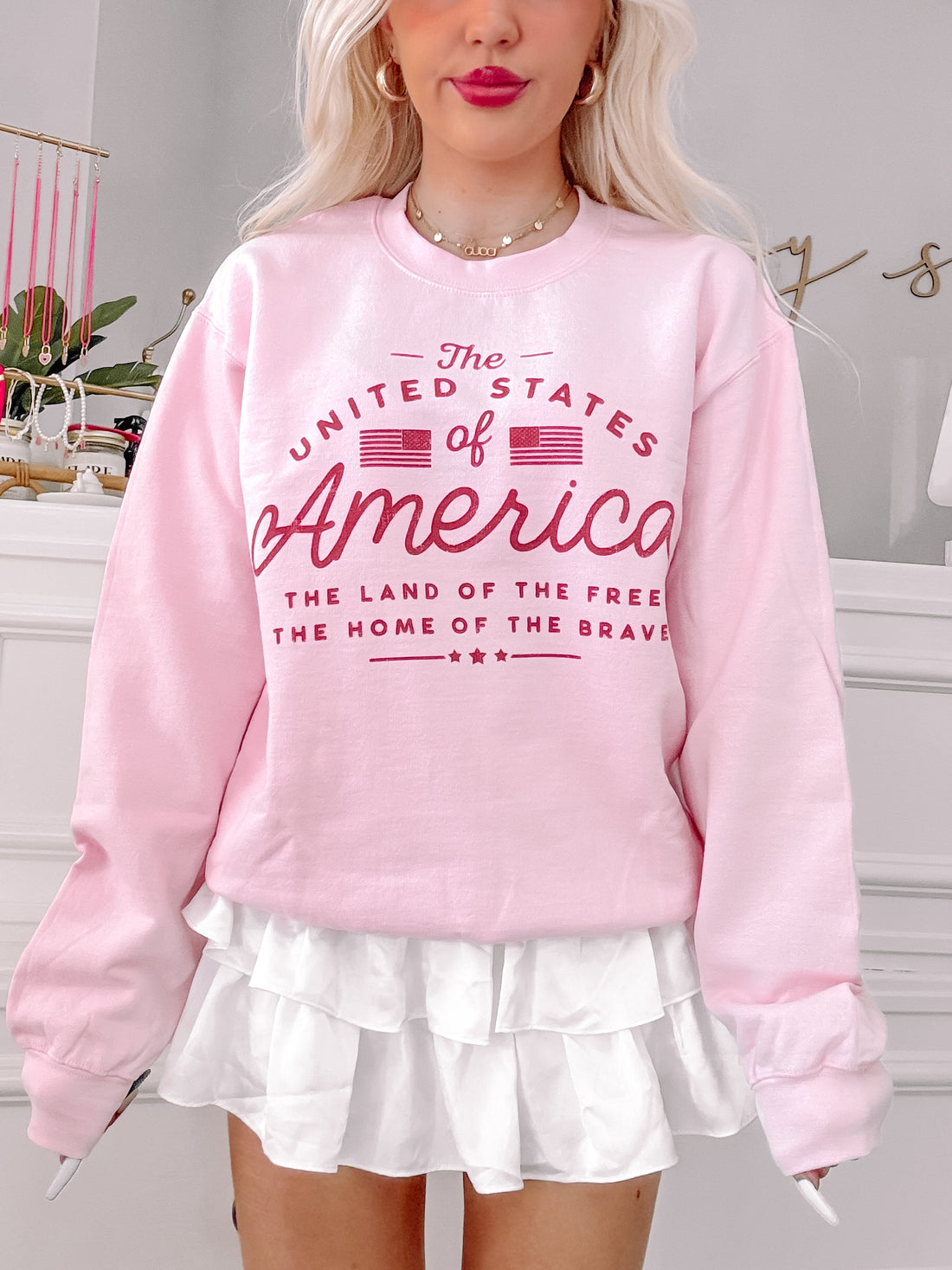 Pink crewneck sweatshirt featuring a patriotic graphic, paired with a white ruffled skirt.
