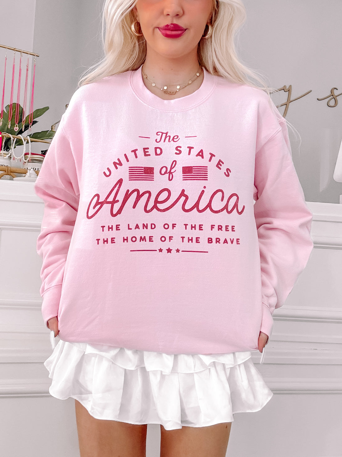 Pink crewneck sweatshirt with patriotic graphic, styled with a white ruffle skirt.
