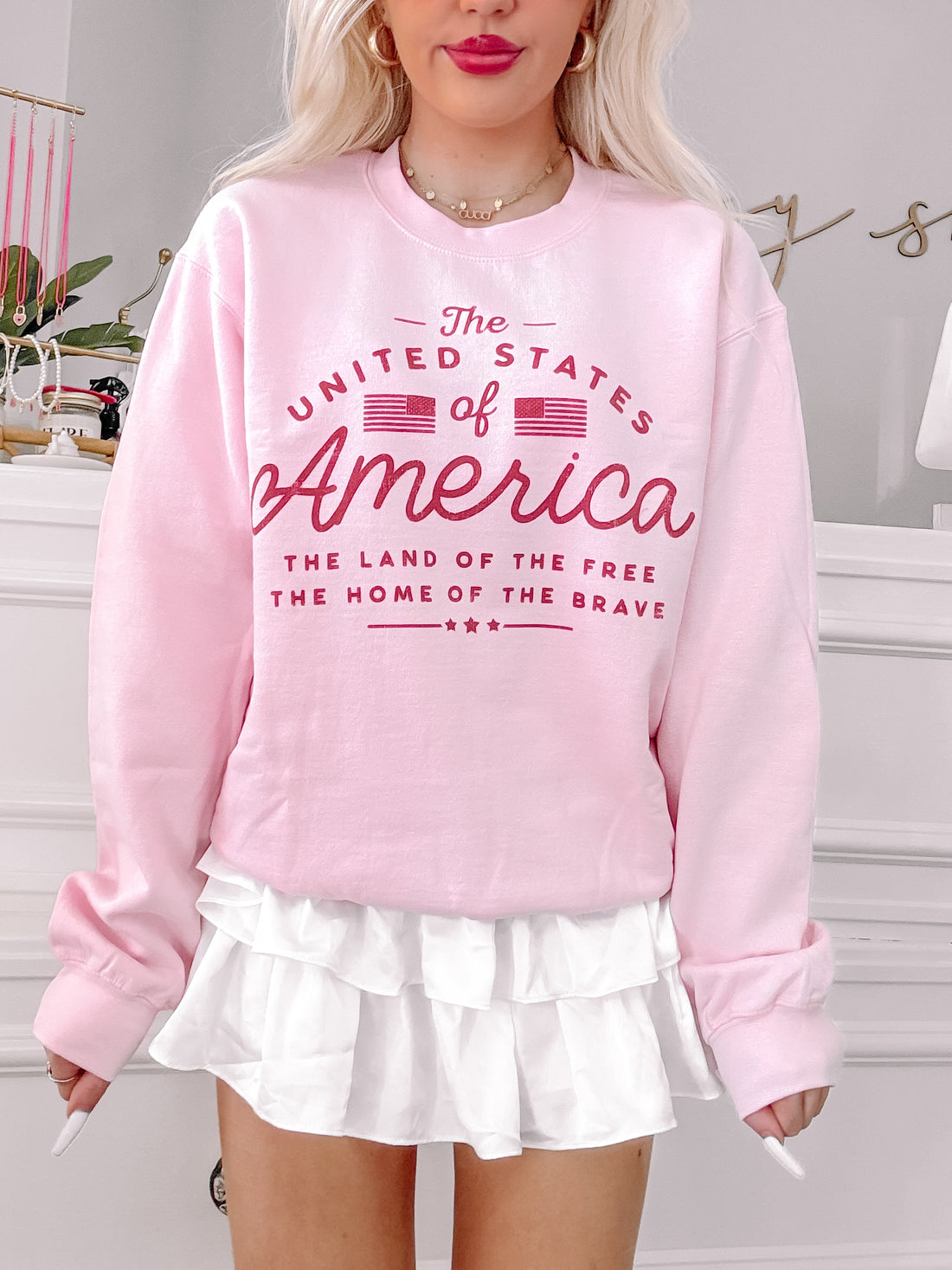 Pink "The United States of America" crewneck sweatshirt with red lettering, featuring an American flag graphic.
