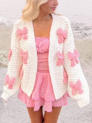 Cream knit cardigan adorned with pink bows, modeled outdoors near water.