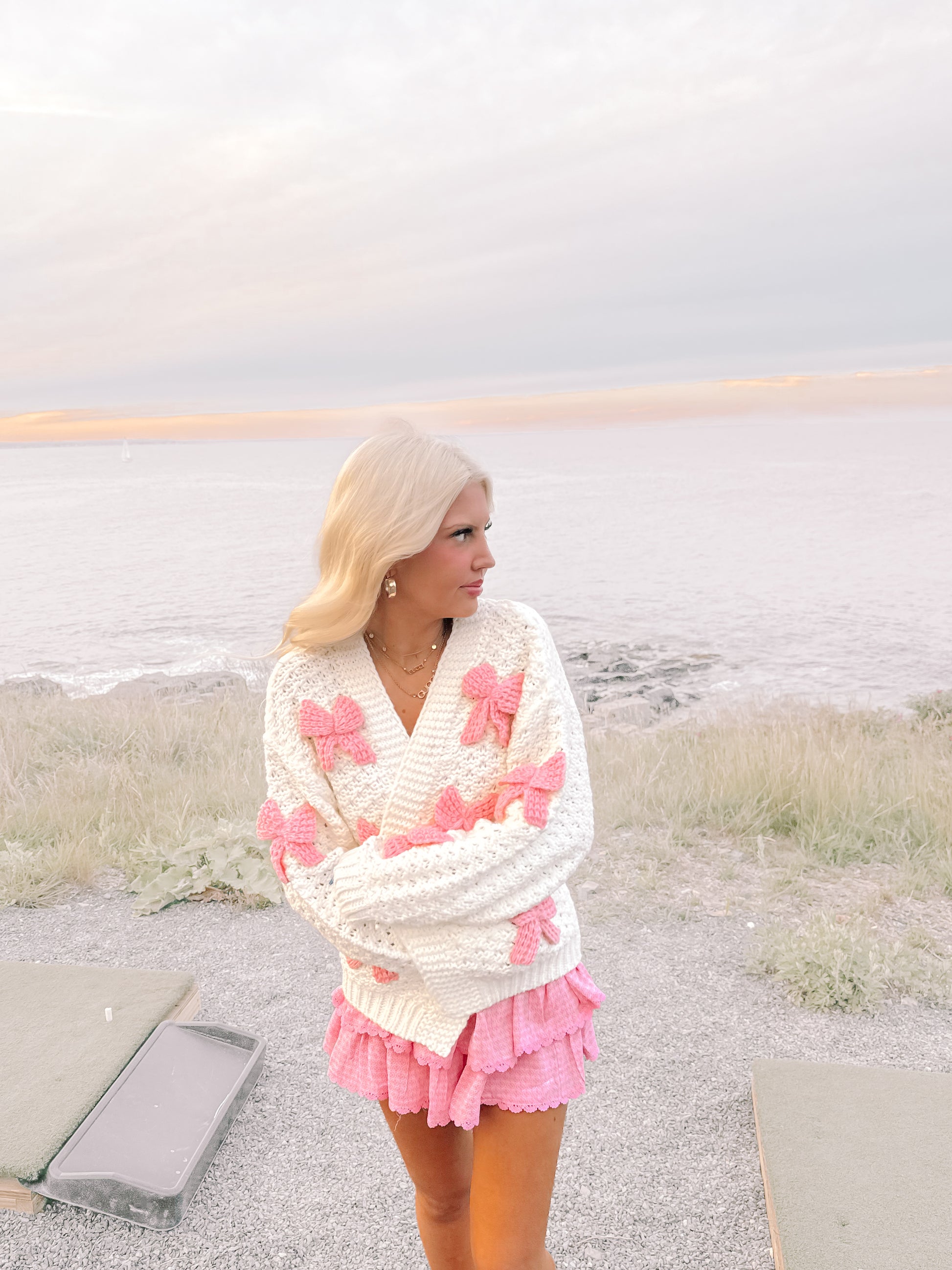 Cream knit cardigan adorned with pink bows, modeled outdoors near the ocean.
