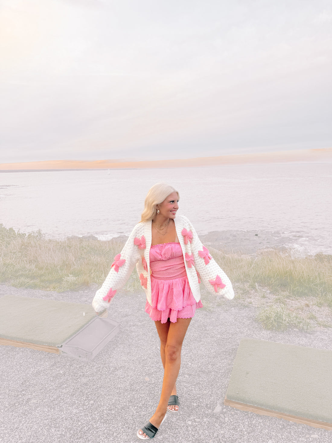 Woman in pink dress and white cardigan adorned with pink bows, posing by the ocean.
