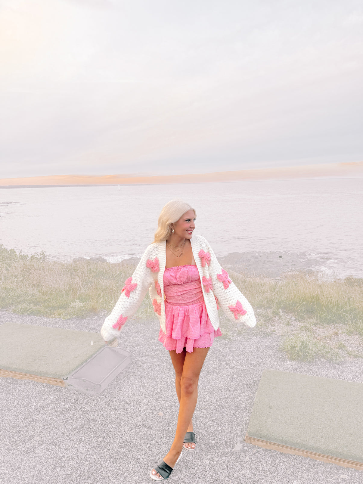 Woman in pink dress and cream knit cardigan with pink bows, seaside setting.
