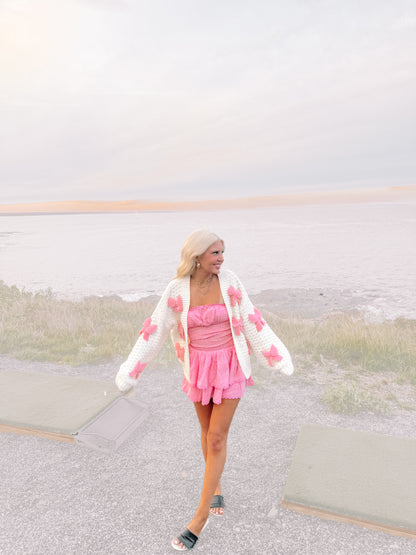 Woman in pink dress and cream knit cardigan with pink bows, seaside setting.
