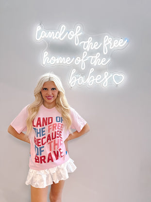 Pink patriotic tee shirt styled with white pleated mini skirt, shown against a wall with a neon sign.
