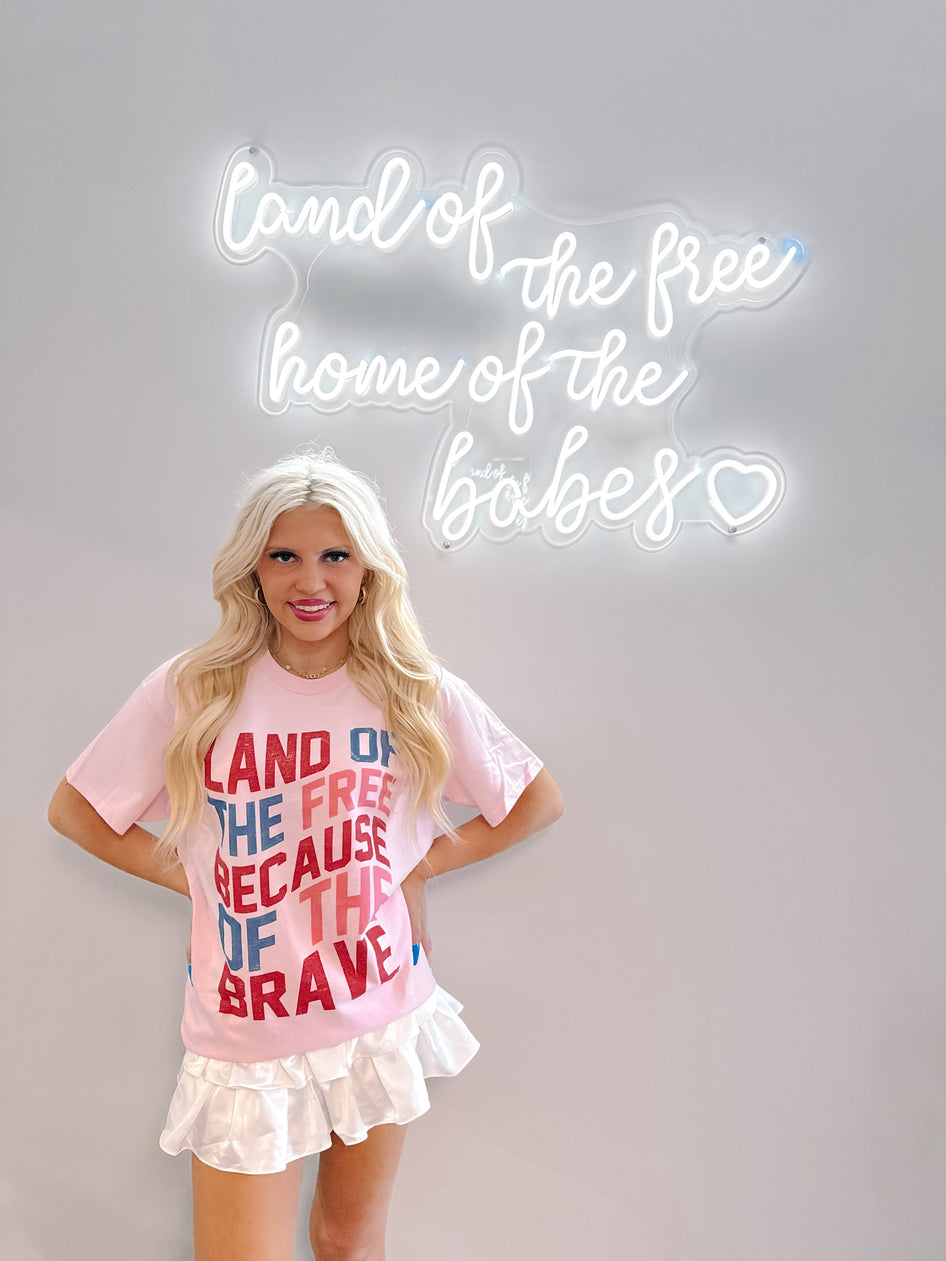 Pink patriotic tee shirt styled with white pleated mini skirt, shown against a wall with a neon sign.
