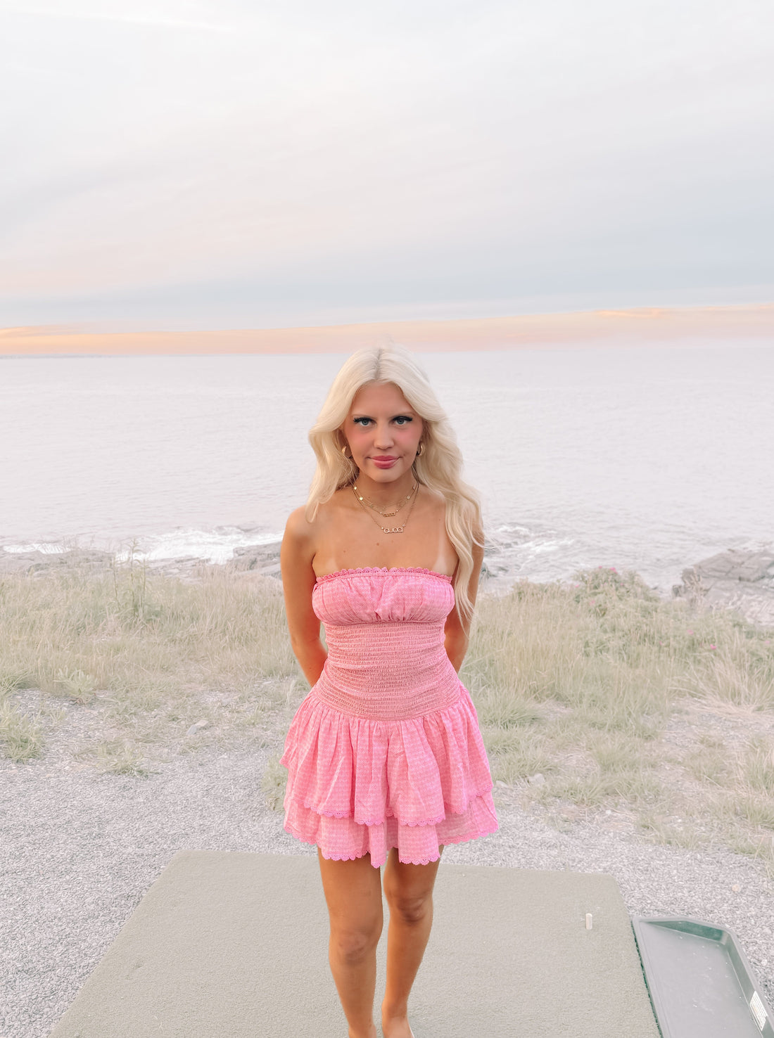 Pink tiered mini dress with eyelet trim, worn outdoors near the ocean at sunset.
