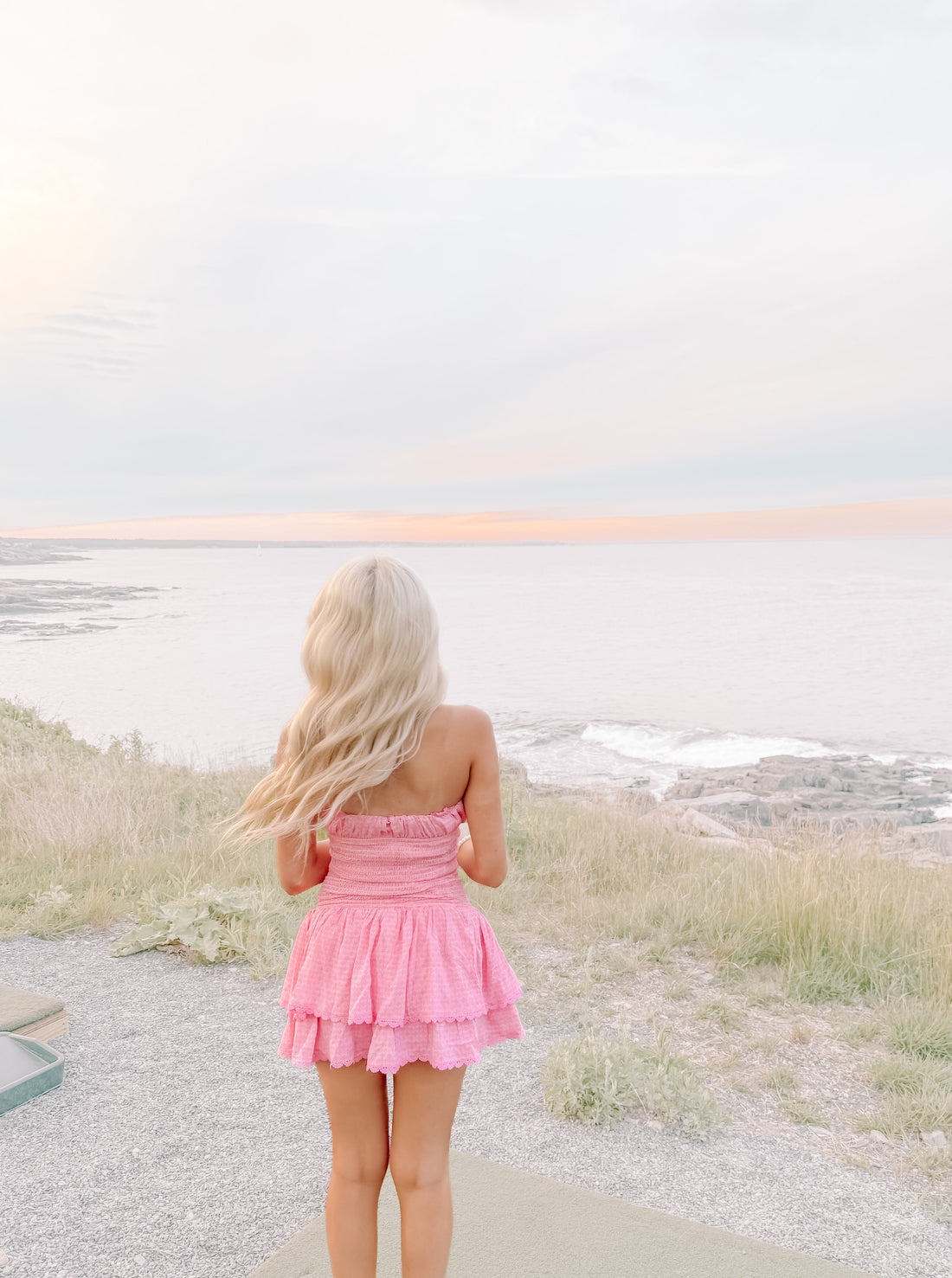 Pink tiered mini dress viewed from behind, overlooking a tranquil ocean at sunset.
