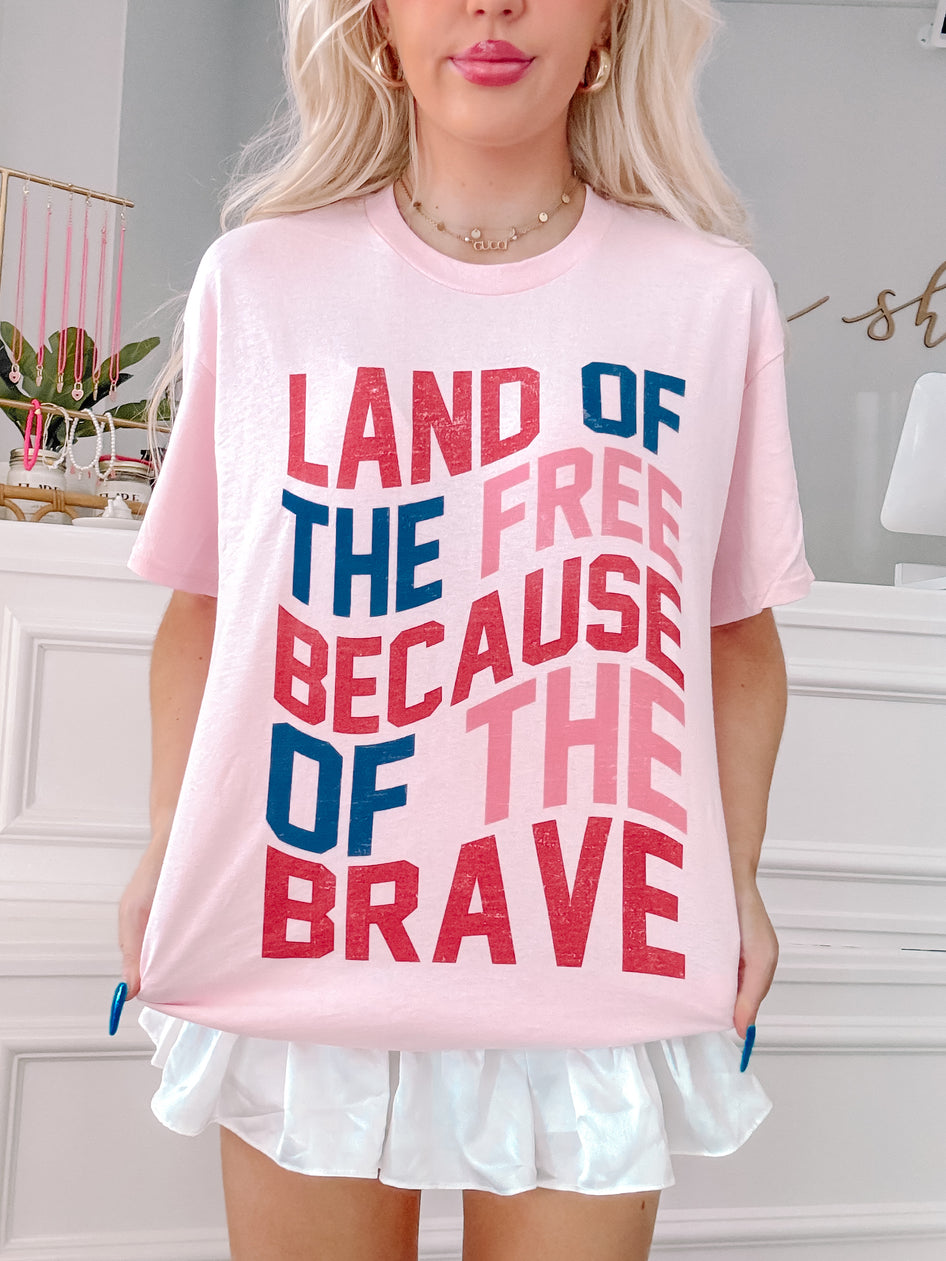 Pink t-shirt displaying "Land of the Free Because of the Brave" in a layered, distressed-style font.
