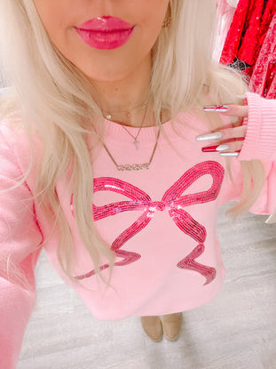 Pink sweater featuring a sparkly pink bow, styled with neutral footwear.

