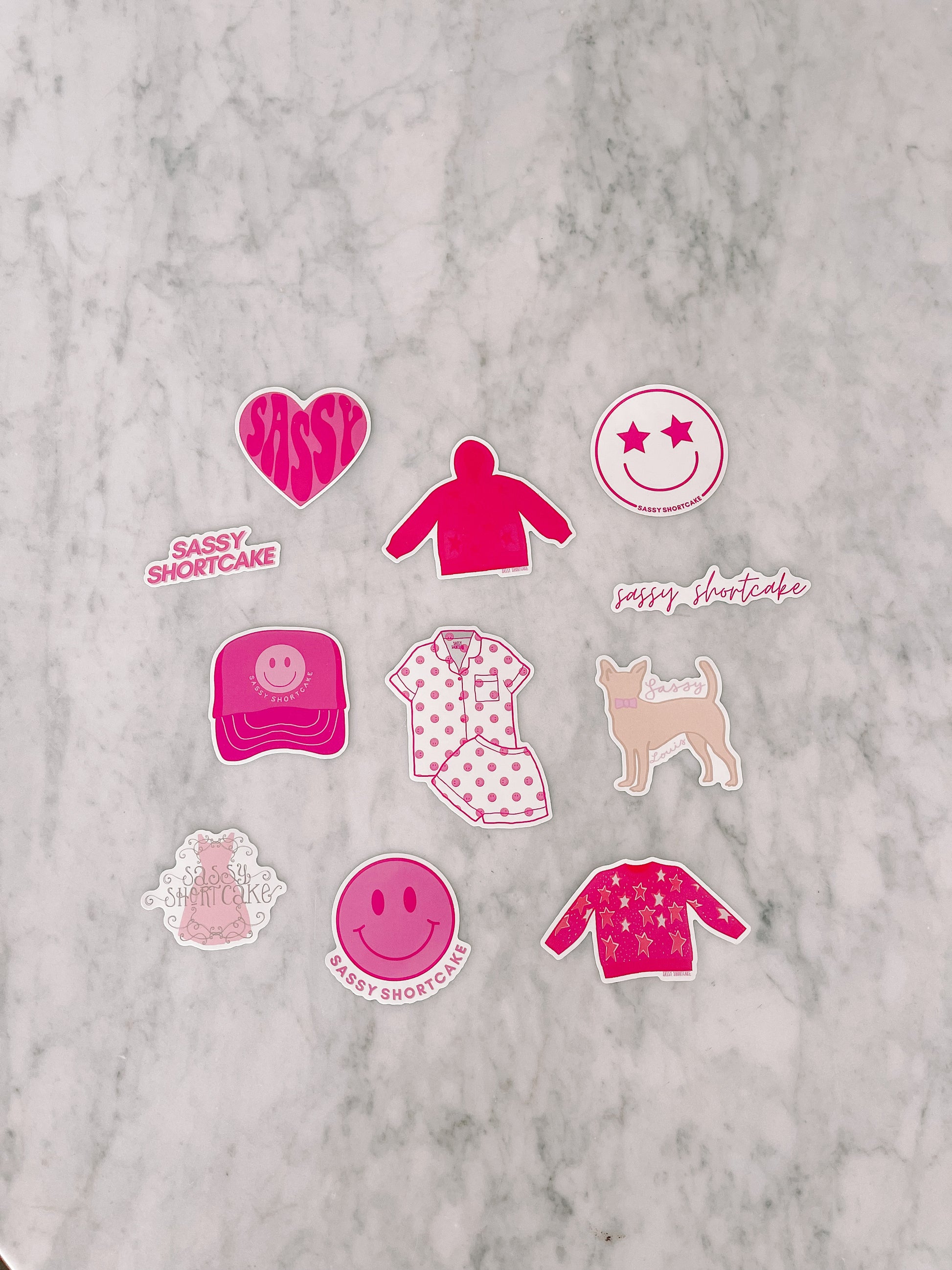 Pink and white sticker assortment on marble; features apparel, pet, and text designs.
