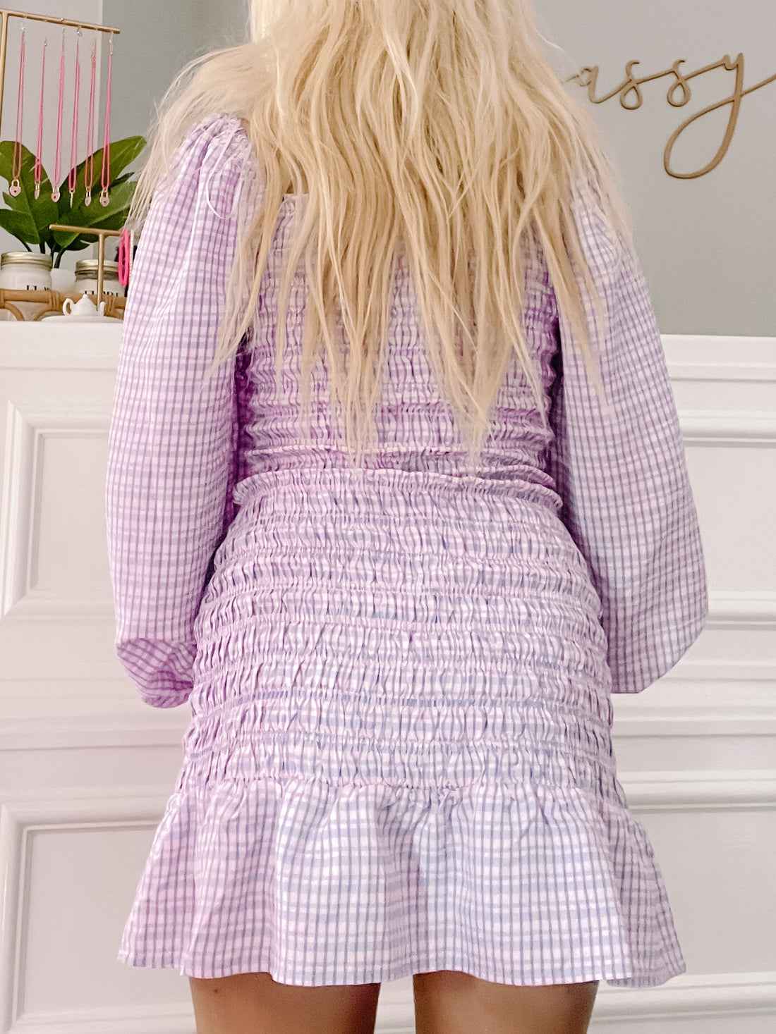 Lilac and white gingham smocked mini dress with long sleeves, shown from the back against a white wall.

