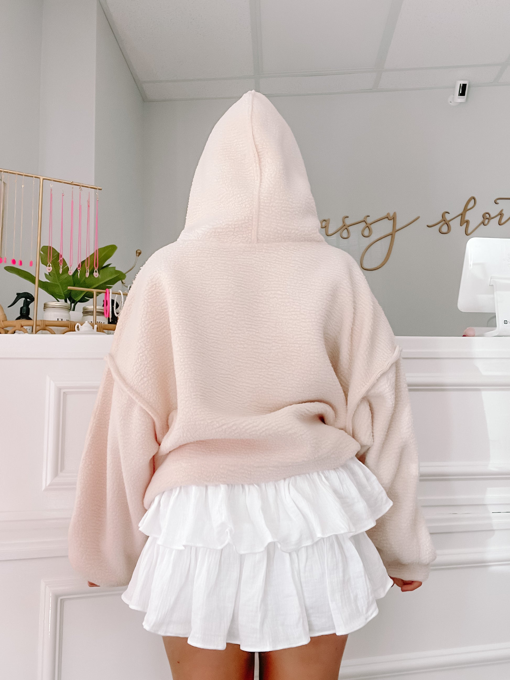 Cream-colored fleece pullover hoodie with a relaxed fit, shown from the back, paired with a white tiered ruffle mini skirt in a boutique setting.
