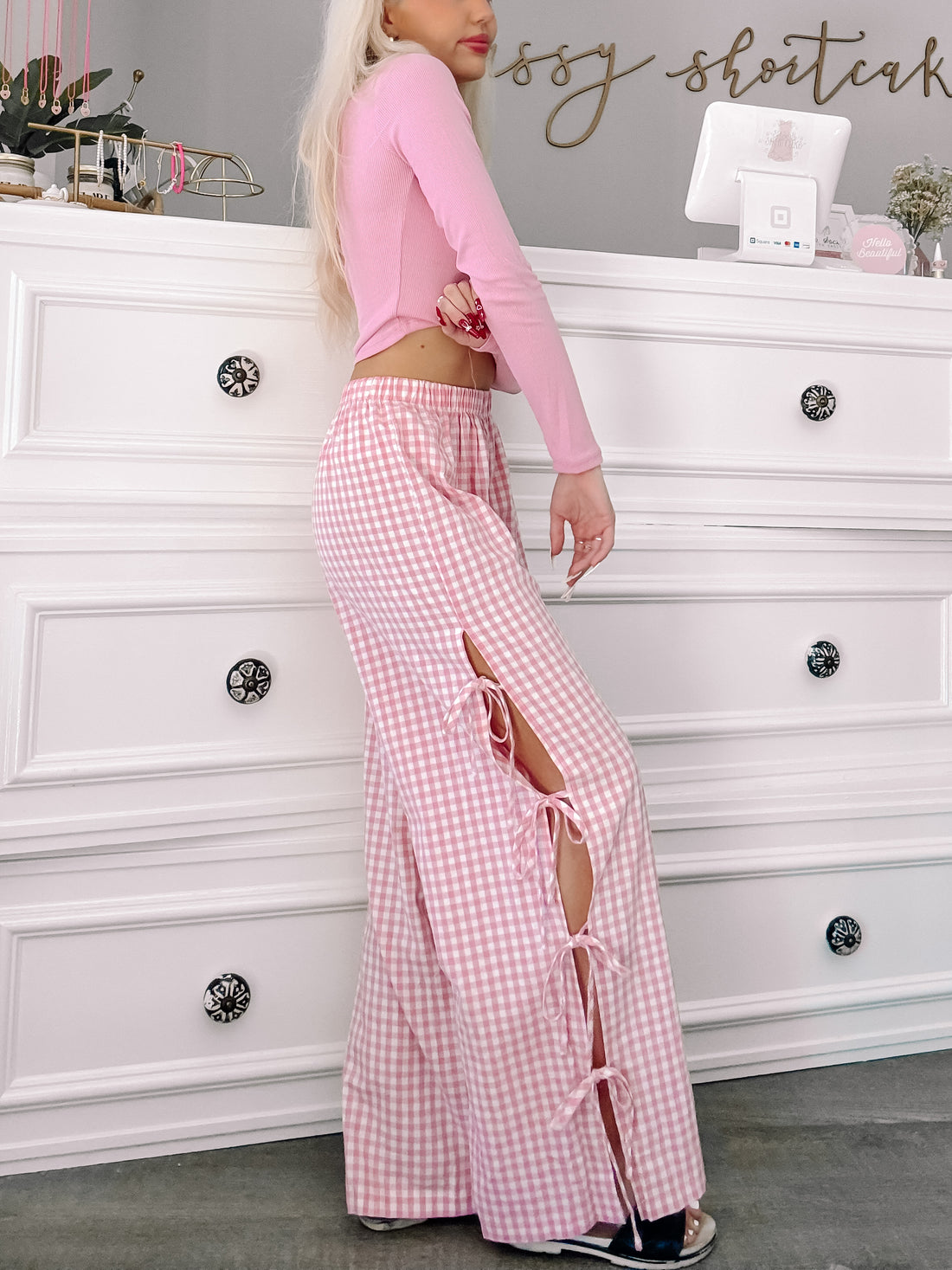 Pink gingham pants with side slits and ties, shown on a model against a white dresser.
