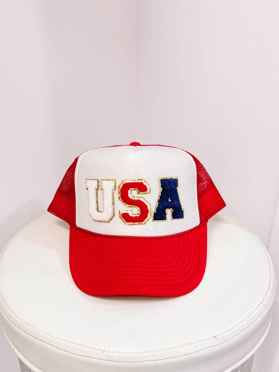 Red, white, and blue USA trucker hat with embroidered lettering on a white stool.
