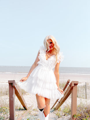 White tiered tulle mini dress styled at the beach; woman in white boots on wooden steps.
